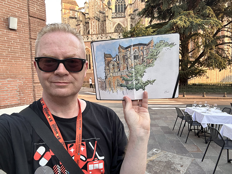 Pete in sunglasses standing in front of Cathedrale St. Etienne in Toulouse holding up a sketch of the cathedral.